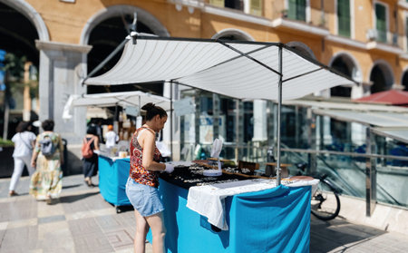 Palma de Mallorca, Spain - Jul 8, 2024: A jewelry vendor organizes items at her market stall in Plaza Mayor, Palma de Mallorca, with people walking through the square in the background on a bright, sunny dayのeditorial素材