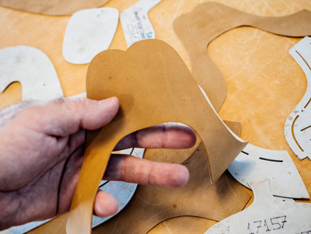 Close-up of a hand holding a leather pattern for shoe production, surrounded by paper templates,...
