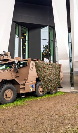 Paris, France - Sep 11, 2024: An armored military vehicle covered with camouflage netting is parked outside a modern glass buildingのeditorial素材