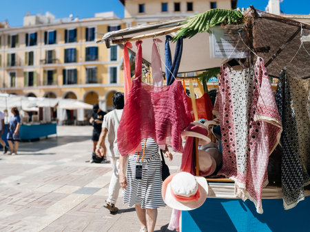 Palma, Spain - Jul 8, 2024: Colorful clothing and hats displayed at an outdoor market stall in Plaza Mayor, with people walking by and historic buildings in the background on a sunny dayのeditorial素材