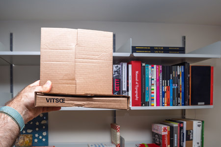 Frankfurt, Germany - Jun 15, 2022: Person holding Vitsoe cardboard packaging box, with a background of colorful bookshelves, showcasing minimalist design and modern interior organizationのeditorial素材