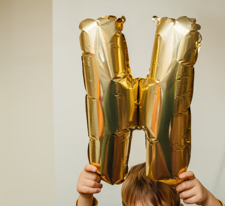 A child holding a shiny gold foil balloon shaped like the letter H against a neutral background, adding a playful and celebratory feel.の写真素材
