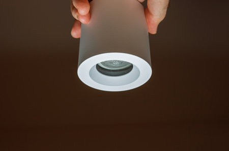 Close-up of a minimalist cylindrical LED light fixture held by a hand, showcasing its modern design and sleek white finish against a dark backgroundの写真素材