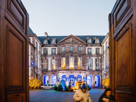Strasbourg, France - Nov 29, 2024: Strasbourg City Hall courtyard decorated for Christmas with festive lights, trees, and visitors passing through a grand wooden doorway at duskのeditorial素材