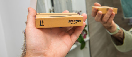Paris, France - Nov 22, 2021: wide image of Hand holding a small Amazon box, showcasing branding and packaging design in a reflective interior setting with additional background elementsのeditorial素材