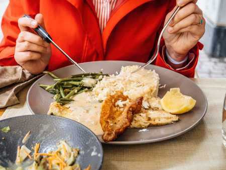 A diner in a red coat using fork and knife to enjoy a fish dish, complemented by lemon and rice, on an outdoor terrace of a restaurantの写真素材