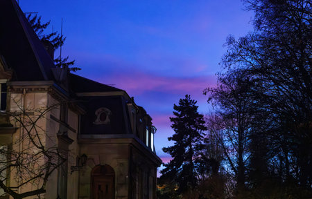 A historic mansion silhouetted against a vibrant twilight sky, with shades of purple and blue blending softly, framed by bare tree branches.の写真素材