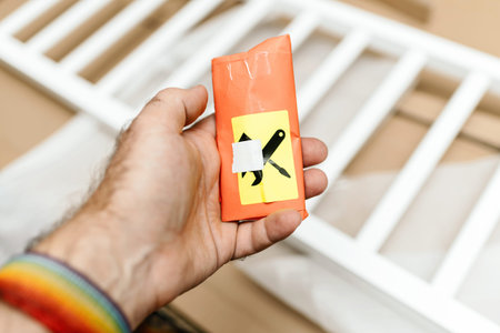 A close-up of a hand holding a small orange packet with a tools icon, indicating assembly tools,...