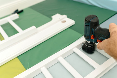 A hand holding a power drill assembling a white wooden frame on a soft green and blue padded surface.の写真素材