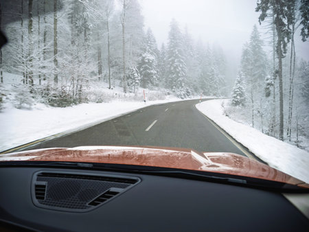 POV A serene winter road in the Black Forest, Germany, bordered by snow-covered trees and a frosty landscape. A car's dashboard is visible in the foreground.の写真素材