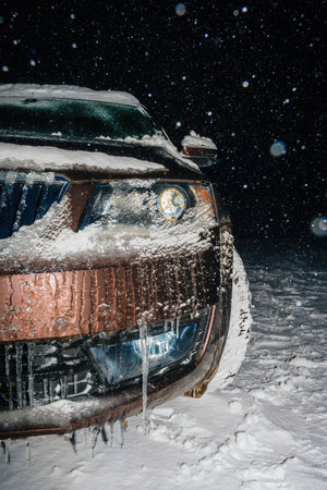 Icy snowflakes accumulate on a car during a blustery winter night, icicles forming beneath the headlights, enhancing the frosty scene.の写真素材