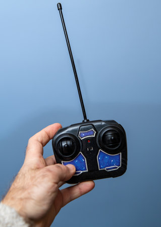 A hand holds a black remote control with a long antenna and blue buttons, designed for operating a toy vehicle or drone, set against a plain light blue backgroundの写真素材