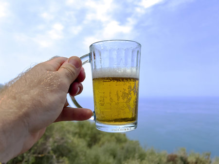A hand holds a glass mug of cold beer against a scenic backdrop of the Mediterranean Sea and clear blue sky, with green coastal vegetation below, suggesting a relaxed moment in Mallorcaの写真素材