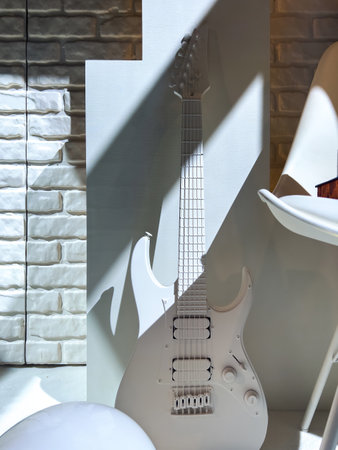 All-white electric guitar with six strings placed against a white backdrop and surrounded by modern white furniture and textured brick wall under diagonal lightの写真素材