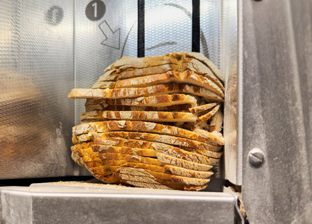 Round loaf of crusty bread partially sliced inside a commercial bread cutting machine, with sharp metal blades and crumbs visible on the stainless steel surfaceの写真素材