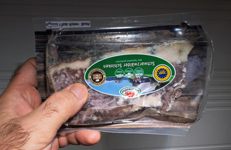 Paris, France - Nov 24, 2023: Hand holding a sealed package of Schwarzwalder Schinken featuring visible layers of cured meat and fat with branded labeling and certification against neutral backgroundのeditorial素材