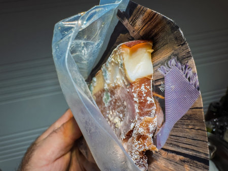 Hand holding an opened vacuum-sealed package containing slices of dry-cured meat with visible salt and fat layers against a wooden-themed printed background and kitchen setting.の写真素材
