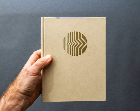 A hand holds a beige hardcover book with a gold embossed circular geometric design on the front...
