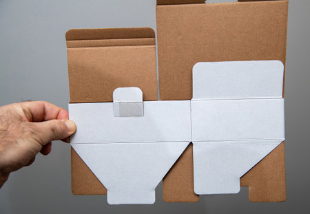 A hand holds a flat, unfolded cardboard box with brown and white surfaces, showing pre-cut flaps, folds, and a glued tab, ready to be assembled for packaging use.の写真素材