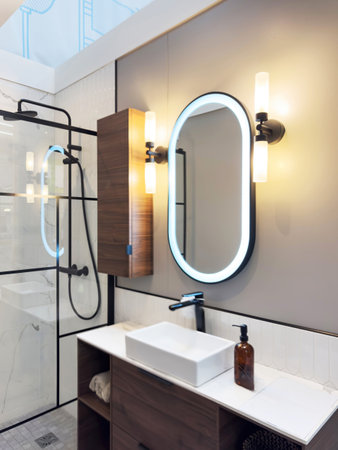 Modern bathroom display featuring a rectangular vessel sink, illuminated oval mirror, wooden vanity with integrated storage, and a glass shower with black framingの写真素材