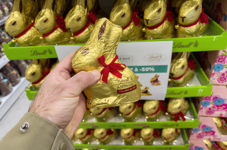 Paris, France- Mar 28, 2025: A hand holds a Lindt Lapin Or milk chocolate bunny wrapped in gold foil with red ribbon in a French supermarket s Easter-themed candy sectionのeditorial素材