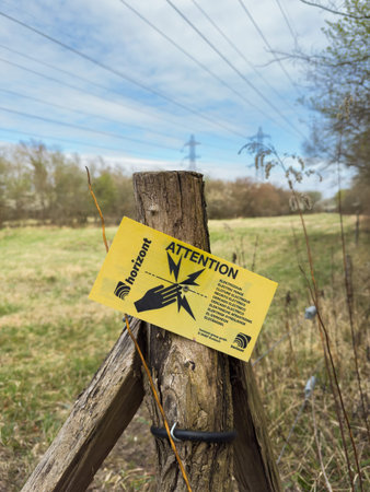 Strasbourg, France - Mar 30, 2025: Yellow multilingual warning sign by Horizont on a wooden post in a grassy field cautions about an electric fence, with power lines and pylons visible in the backgroundのeditorial素材
