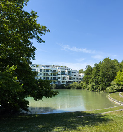 A luxurious modern residential complex in France stands beside a tranquil green water pond surrounded by lush trees under a bright blue summer skyの写真素材