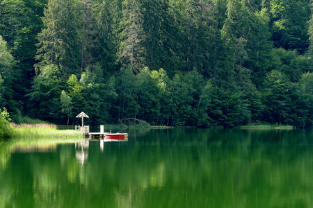 Lake in the mountains. Around surrounded by trees. A marina with an umbrella and a red boat. Reflection of trees and boats in the water. The water is calm, without waves. An atmosphere of peace and relaxation.の写真素材