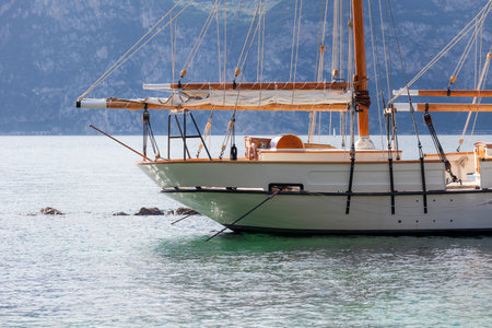 Front and center, a moored sailboat rests in calm waters, its wooden mast and taut rigging framed...