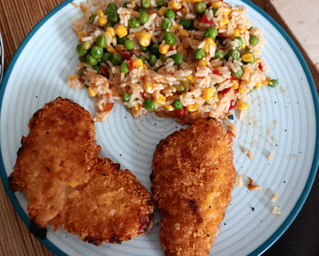 Breaded chicken with rice and vegetables (green peas, red peppers and corn) on a blue plateの写真素材