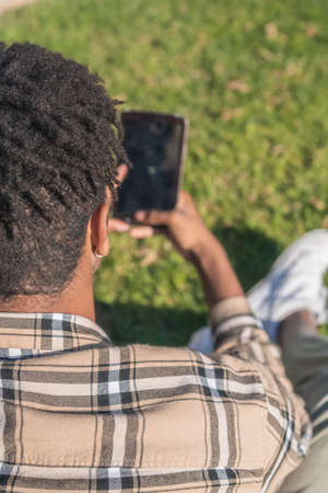 black afro american man with tablet in hand beige plaid jacket in a park sitting on a green grass warm colors in old townの写真素材