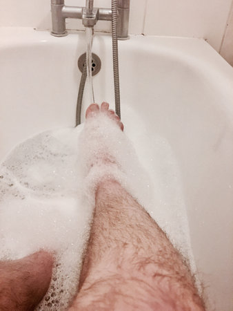 Man getting the bath. Water on red hairy leg in the bathroom.