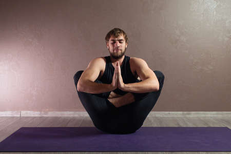 Young sporty man practicing yoga, meditating in Half Lotus pose, working out, wearing sportswear, indoor full length, brown yoga studioの写真素材