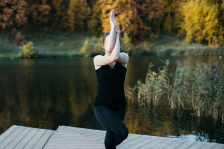 Young woman doing yoga asana in the nature with the lake view. Evening workout outdoors, sports and healthy lifestyleの写真素材