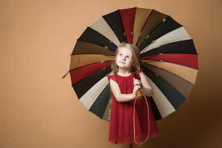 Happy funny child with beautiful umbrella posing on orange wall background. Girl is wearing red dress.Rainの写真素材