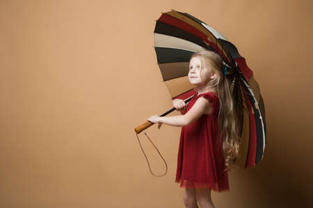 Happy funny child with beautiful umbrella posing on orange wall background. Girl is wearing red dress. Rainの写真素材