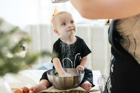 A young mother in an apron and curlers is preparing a birthday cake in the kitchen with her little child. Bright and cozy kitchenの写真素材