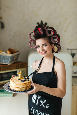 A young mother in an apron and curlers is preparing a birthday cake in the kitchen with her little...