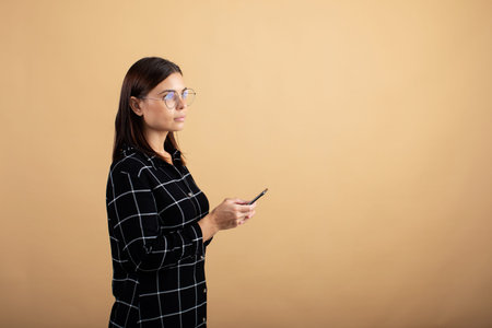 A young woman in a plaid dress stands on an orange background and plays with her phoneの写真素材