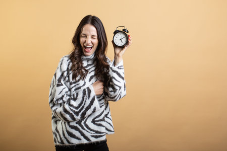 Image of a young beautiful woman posing isolated against an orange wall holding an alarm clock