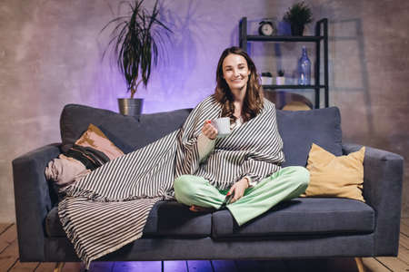 A young woman sits on a sofa in a cozy room wrapped in a blanket, drinking tea and watching TV.の写真素材