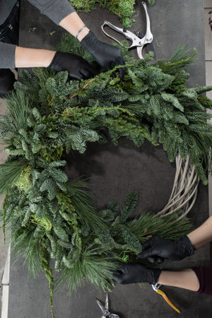 Top view of florist hands making Christmas wreath with fir branches on the table.
