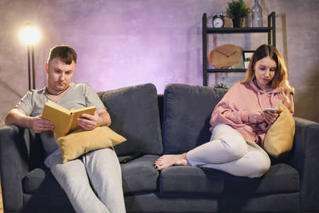 A man reading a book, a woman texting. Man and woman sitting in a cozy apartment on the couchの写真素材