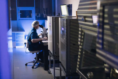 In a Modern Laboratory Scientist Conduct Experiments. Chief Research Scientist Adjusts Specimen in a Petri Dish and Looks on it Into Microscopeの写真素材