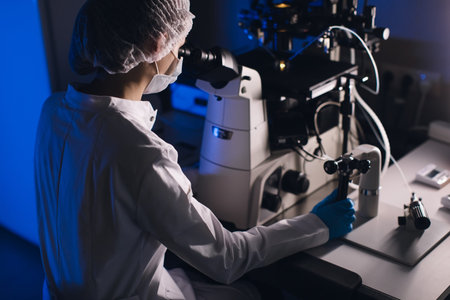 In a Modern Laboratory Scientist Conduct Experiments. Chief Research Scientist Adjusts Specimen in a Petri Dish and Looks on it Into Microscopeの写真素材
