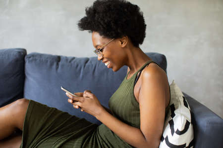 Happy african American young woman sit relax on cozy couch happy to move to new apartment. Smiling black girl rest on comfortable sofa in living room dreamingの写真素材