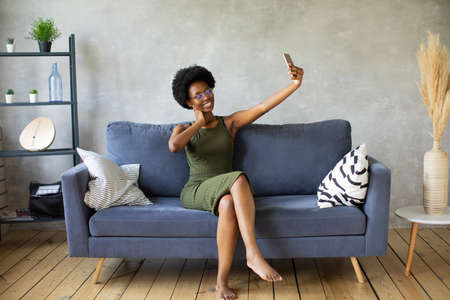 Happy african American young woman sit relax on cozy couch happy to move to new apartment. Smiling black girl rest on comfortable sofa in living room dreamingの写真素材