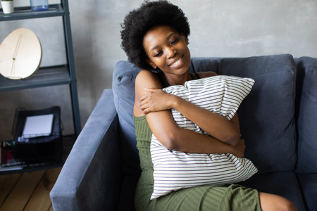 Happy african American young woman sit relax on cozy couch and hugs a pillow. Happy to move to new...