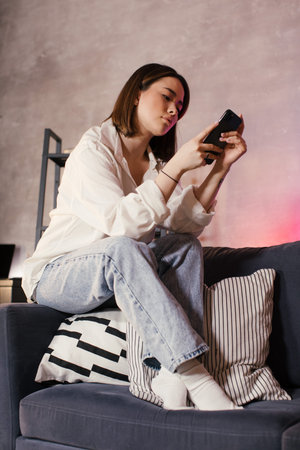 Young asian woman sitting on a sofa in a cozy room looks at the phone
