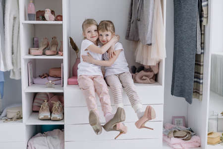 Family wardrobe. Happy twin sisters on shelve in closet with natural organic clothes in cozy dressing room play joyfullyの写真素材